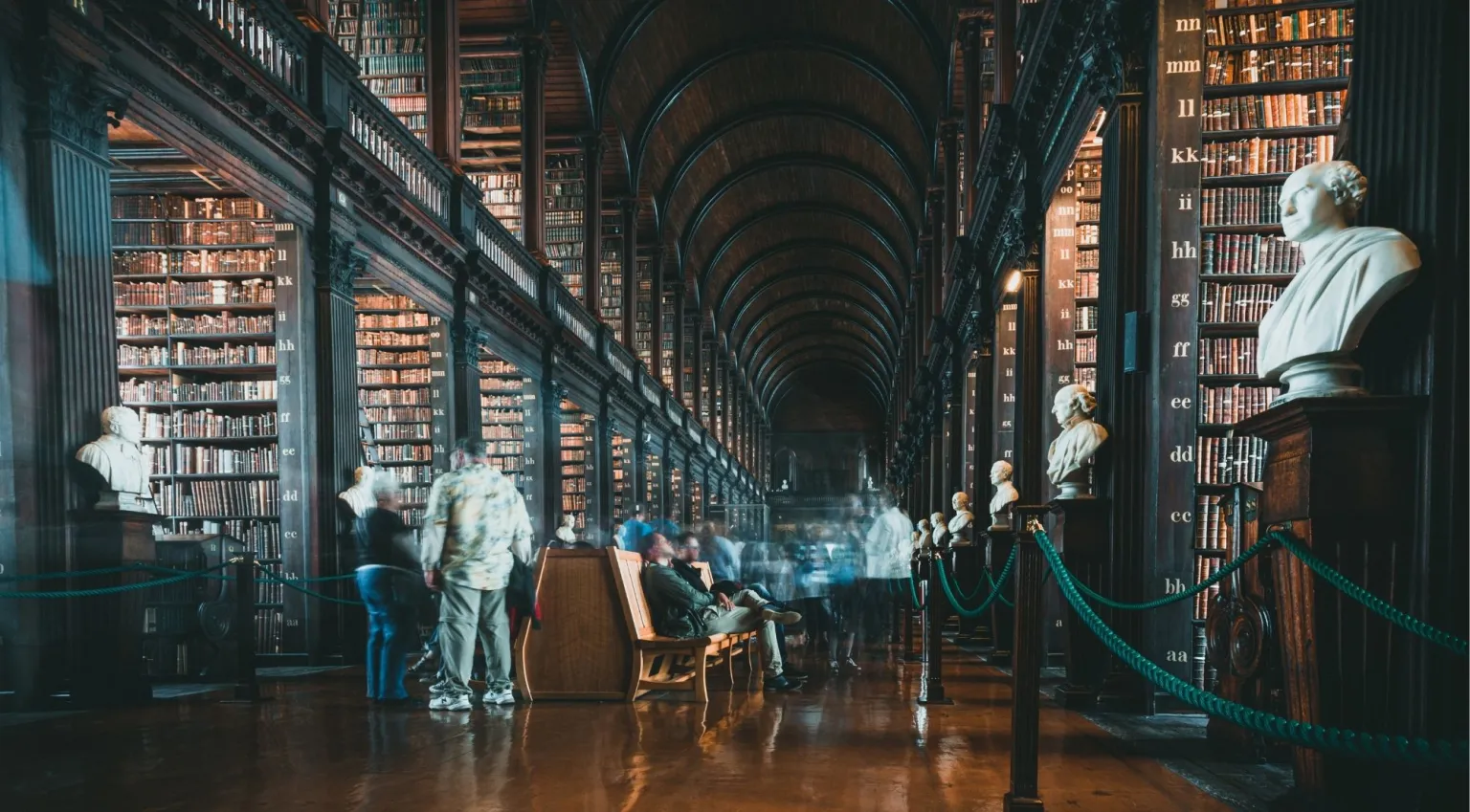 Visitors exploring the Long Room beneath the barrel-vault ceiling, with marble busts and display cases